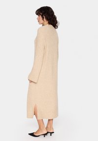 Abito in maglia beige con maniche lunghe, spacco laterale sull'orlo, texture morbida e vestibilità ampia. Indossato con mule nere con tacco.