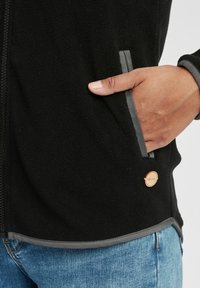 Mustmustööst must fleece-jakk hallide aktsentidega, tõmblukuga ees, küljetaskud ja pronksist logo detail. Sile tekstuur ja vabalt istuv lõige.