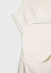Bílá texturovaná tkanina s krabatým designem. Má široký střih a zaoblené otvory po stranách. Jednoduchý a minimalisticý styl.
