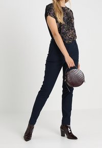 Top floral negro con mangas cortas, pantalones ajustados azul marino, botines burdeos brillantes y un bolso de mano redondo con textura.