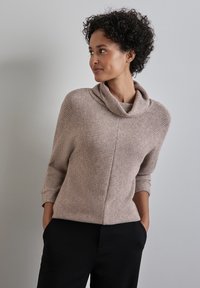 Maglione lavorato a maglia in taupe chiaro con scollo a caracoll, maniche tre quarti e un tessuto strutturato. Abbinato a pantaloni neri.