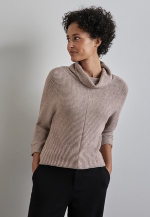 Striktrøje i lys taupe med cowl-halsudskæring, trekvartærmer og et tekstureret stof. Kombineret med sorte bukser.