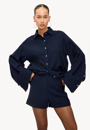 Chemise bleu marine surdimensionnée à boutons avec des manches larges et une poche poitrine unique, complétée par un short assorti ; des boutons argentés soulignent les poignets.
