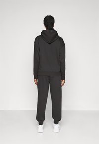 Schwarze Kapuzenjacke mit großzügiger Kapuze und herunterhängenden Schultern, kombiniert mit schwarzen Jogginghosen; beide aus einem weichen, strukturierten Stoff gefertigt.