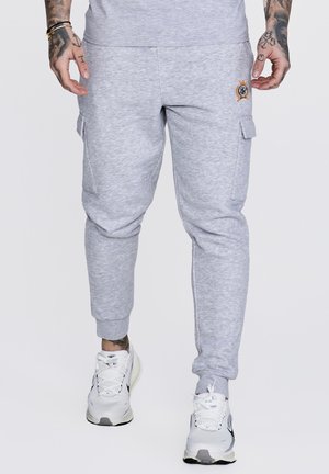 Lysegrå joggebukser med sidelommer og en liten brodert logo på høyre lår, brukt med hvite joggesko og tatoverte hender.