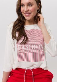 Femme souriante, portant un haut à manches longues blanc avec des lignes verticales rouges formant le mot "FASHION" et un pantalon rouge avec cordon de serrage.