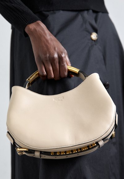 Main tenant un sac à main en cuir beige de Moschino avec une poignée en métal doré et une bandoulière noire sur une jupe noire à boutons.