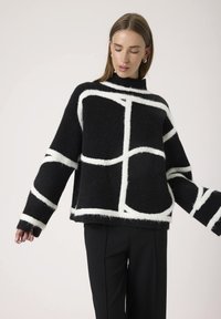 Mujer vistiendo un suéter negro oversize con gruesas líneas geométricas blancas y pantalones negros, de pie con los brazos ligeramente extendidos.