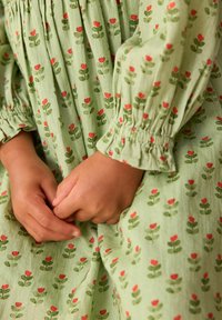 Grünes Kleid mit floralem Muster, das rote Blumen und grüne Blätter zeigt. Geschopfte Ärmel und strukturierter Stoff sind sichtbar, Hände am Bauch verschränkt.