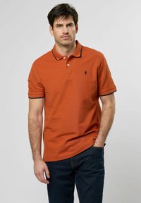 Polo majica boje rđe s ovratnikom, kratkim rukavima i mornarsko plavim rubovima. Ima mali grb na prsima i teksturirani materijal.