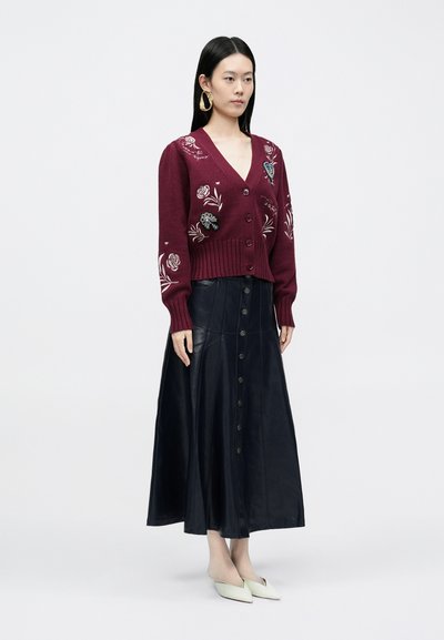 Bordowy sweter kardigan z dzianiny z kwiatową haftem, zestawiony z czarną skórzaną spódnicą na guziki oraz jasnymi mulesami.