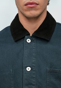 Chupa de denim azul oscuro con cuello de pana negro, que presenta un botón a presión plateado, bolsillo en el pecho y tejido texturizado.
