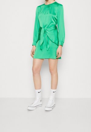 Mujer con un mini vestido de satén verde brillante de manga larga con detalle de cintura atada, calcetas blancas de Nike y zapatillas blancas con plataforma.
