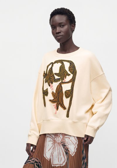 Γυναίκα που φοράει εκρού oversized φούτερ με καφέ και ροζ κεντήματα λουλουδιών και καφέ πλισέ φούστα με μεγάλα λουλουδένια σχέδια.