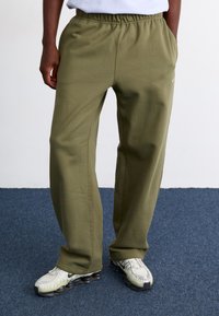 Calças de treino verde-azeitado com uma cintura elástica, bolsos laterais e detalhes de costura visíveis. Usadas com sapatilhas de tonalidade clara.