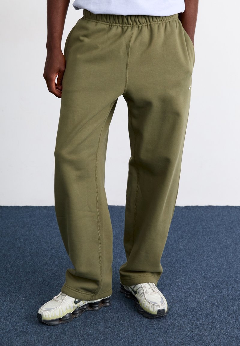 Calças de treino verde-azeitado com uma cintura elástica, bolsos laterais e detalhes de costura visíveis. Usadas com sapatilhas de tonalidade clara.