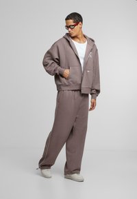 Hooded zip-up sweatshirt and matching sweatpants in light brown. Sweatshirt has a front pocket; pants feature side stripes and elastic waistband.