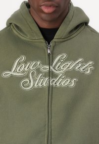 Haut à capuche zippé vert olive avec un logo brodé "Low Lights Studios" en vert clair. Doté d'une capuche et d'un tissu doux et texturé.