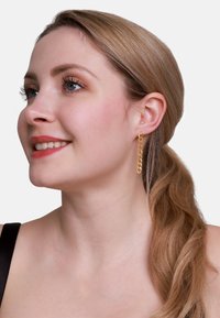 Orecchini a pendente in oro con forme ovali collegate, caratterizzati da una finitura lucida e un design leggero, indossati da una persona con capelli castano chiaro.