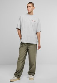 Tricou supradimensionat de culoare gri deschis, cu textul "INTERZIS" în roșu pe piept, asociat cu pantaloni largi de culoare verde măsliniu și adidași deschiși.