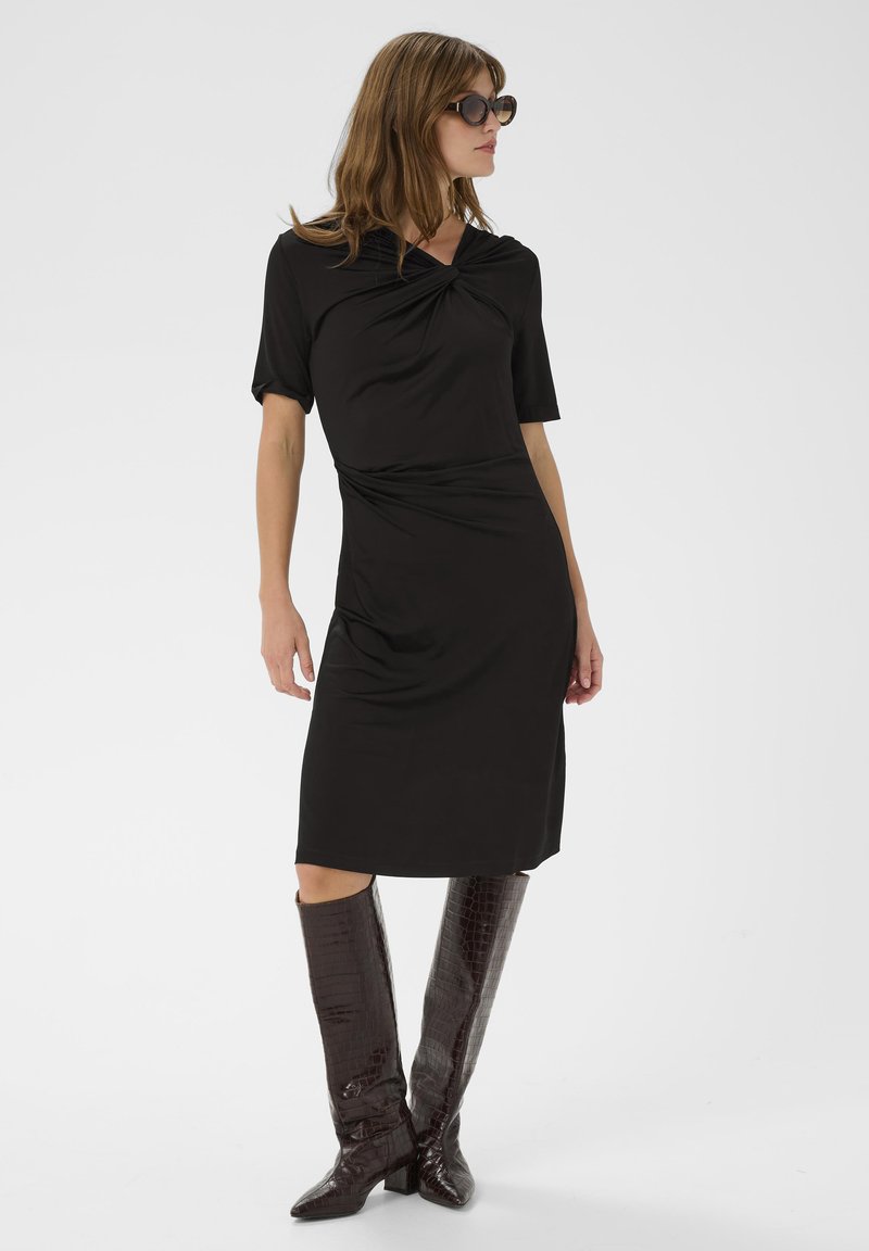 Mujer con un vestido negro de longitud a la rodilla y cuello retorcido, botas oscuras de cocodrilo hasta la rodilla y gafas de sol redondas, de pie sobre un fondo blanco.