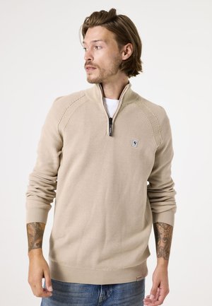 Beige gebreide pullover met een halve rits kraag, geribde manchetten en zoom, en een klein logo patch op de borst. Heeft een textuur stof en een relaxte pasvorm.