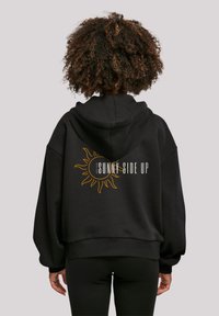 Black oversized hoodie featuring a sun graphic and "SUNNY SIDE UP" text in yellow on the back. Soft material with a relaxed fit.