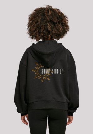 Black oversized hoodie featuring a sun graphic and "SUNNY SIDE UP" text in yellow on the back. Soft material with a relaxed fit.