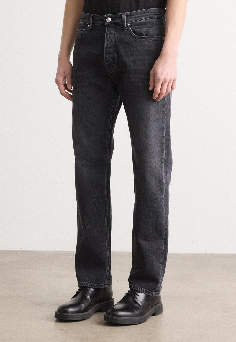 Jean en denim foncé à coupe droite, avec un design classique à cinq poches, un léger délavage et une taille mi-haute. Porté avec des chaussures en cuir noires.