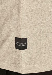 Világosszürke, puha anyagú póló, hajtogatott fekete címkével, rajta fehér szöveggel: "ROCAWEAR" és "ESTABLISHED 1999".