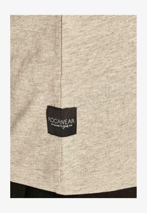 Világosszürke, puha anyagú póló, hajtogatott fekete címkével, rajta fehér szöveggel: "ROCAWEAR" és "ESTABLISHED 1999".