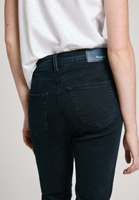 Detailní záběr osoby v tmavě modrých džínách s zadními kapsami a bílou košilí zasunutou do kalhot, s viditelnou částí paží a trupu.