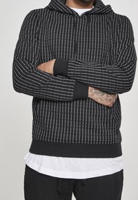 Hombre con mano tatuada, vistiendo una sudadera negra con rayas verticales blancas y una pulsera plateada, con los brazos cruzados sobre una camisa blanca y pantalones negros.