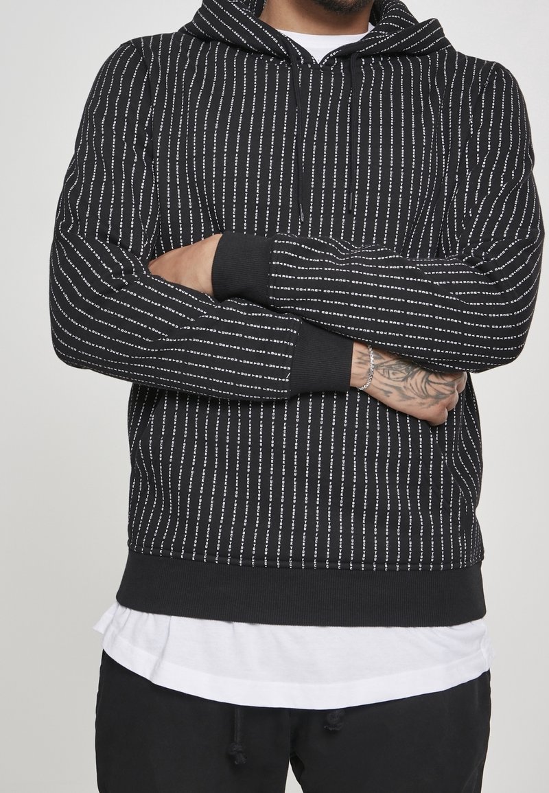 Hombre con mano tatuada, vistiendo una sudadera negra con rayas verticales blancas y una pulsera plateada, con los brazos cruzados sobre una camisa blanca y pantalones negros.