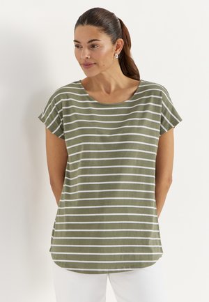 Ruskeahiuksinen nainen poninhännällä, pukeutunut vihreävalkoiseen raidalliseen lyhythihaiseen paitaan ja valkoisiin housuihin, katsoo sivulle.