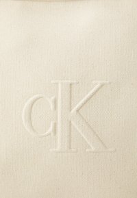 Beige látková taška s texturovaným povrchem, s vyraženým logem "cK". Zaměřte se na jemné zrno a minimalistický design.