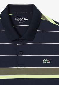 Polo shirt blu navy con righe bianche orizzontali, caratterizzato da un logo Lacoste verde e un accento verde lime angolato sul colletto. Vestibilità regolare.