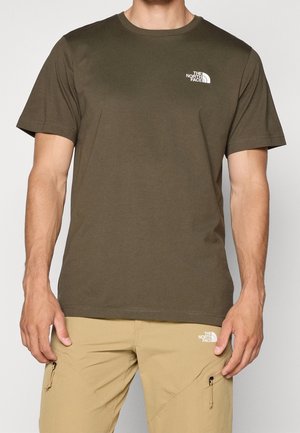 Mężczyzna ubrany w oliwkowozielony t-shirt z krótkim rękawem oraz beżowe spodnie, oba z białymi logo The North Face na piersi i udzie.