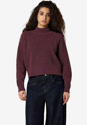 Giovane donna con capelli lunghi e ricci che indossa un maglione a coste color bordeaux e jeans blu scuro a vita alta, in piedi davanti a uno sfondo semplice.