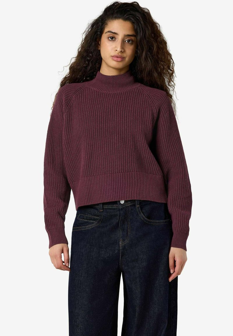 Jeune femme aux longs cheveux bouclés, vêtue d'un pull côtelé bordeaux et d'un jean taille haute bleu foncé, debout devant un fond uni.