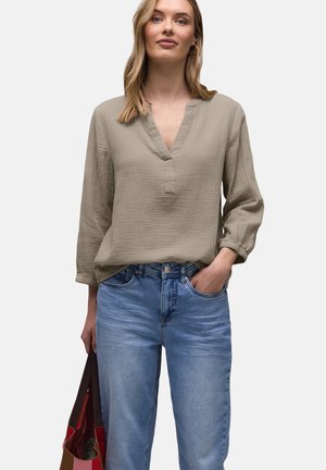Frau trägt eine beige langärmlige V-Ausschnitt-Bluse und blaue Jeans, hält eine gemusterte Tragetasche und hat eine Hand in der Tasche.