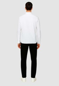 Camicia bianca a maniche lunghe con polsini abbottonati, indossata con pantaloni neri. La camicia presenta un design dritto sul retro e un orlo leggermente curvato.