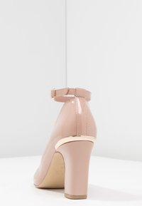 Nude lacklädershögklackad sko med ankelrem och guldaccent på klacken, med rund tå och glansig yta.