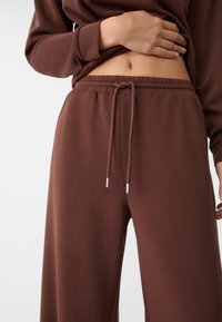 Bruna loungebyxor tillverkade av mjukt tyg, med elastisk midja med dragsko och silverfärgade metalldetaljer för justering.
