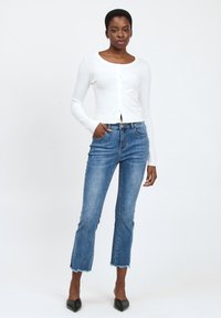 Cardigan blanco, recortado, con mangas largas y botones en la parte delantera, combinado con unos jeans de mezclilla azules de tiro alto, con dobladillos deshilachados y un corte ajustado.
