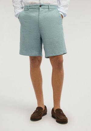 Mann, der hellblaue maßgeschneiderte Shorts mit Vordertaschen, ein eingestecktes gestreiftes Langarmhemd und braune Wildleder-Loafer trägt und steht.
