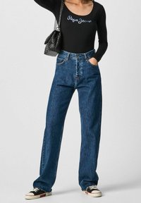 Schwarzer Langarm-Bodysuit mit dem Text 'Paige Jeans', kombiniert mit blauen hoch taillierten, weit geschnittenen Jeans. Accessoires: eine kleine schwarze Handtasche und Sneakers.
