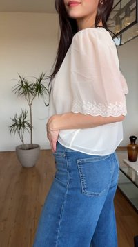 Žena s dugom tamnom kosom nosi prozirnu bijelu bluzu s čipkastim rukavima i plave traperice, stoji u sobi s drvenim podom i sobnom biljkom u tegli.