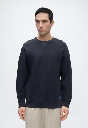 Homme aux cheveux noirs courts portant un sweat-shirt bleu foncé à manches longues et un pantalon beige, debout devant un fond uni.