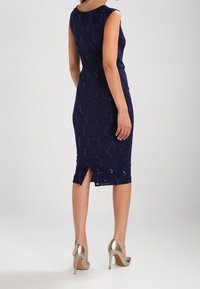 Tyrkysově modré bezramínkové šaty s těsným střihem, s flitrovou texturou, délky ke kolenům a rozparkem na zadní části. Nosené s metalickými podpatky.